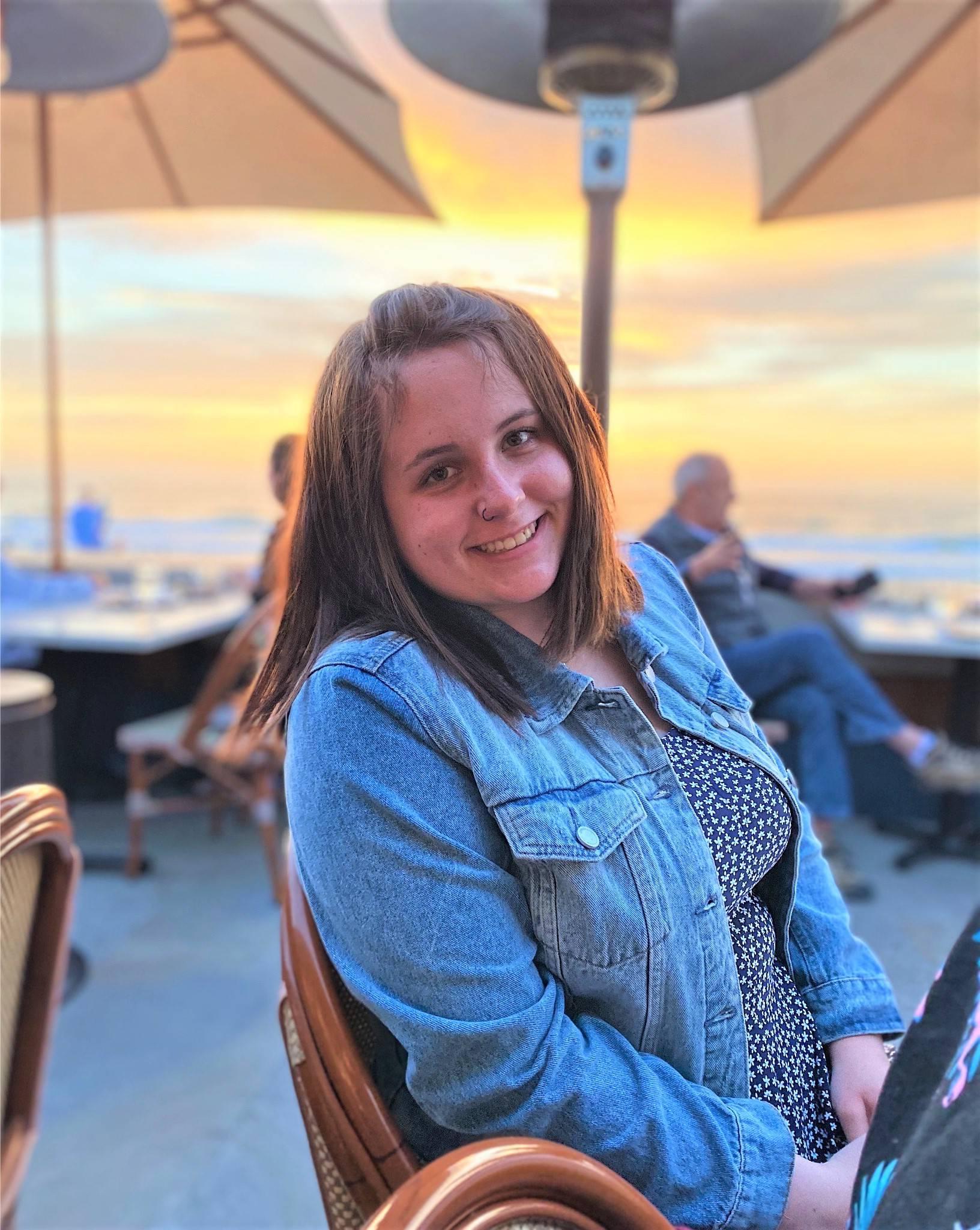 Woman with shoulder length brown hair wearing jean jacket by a table with a sunset beach in the background